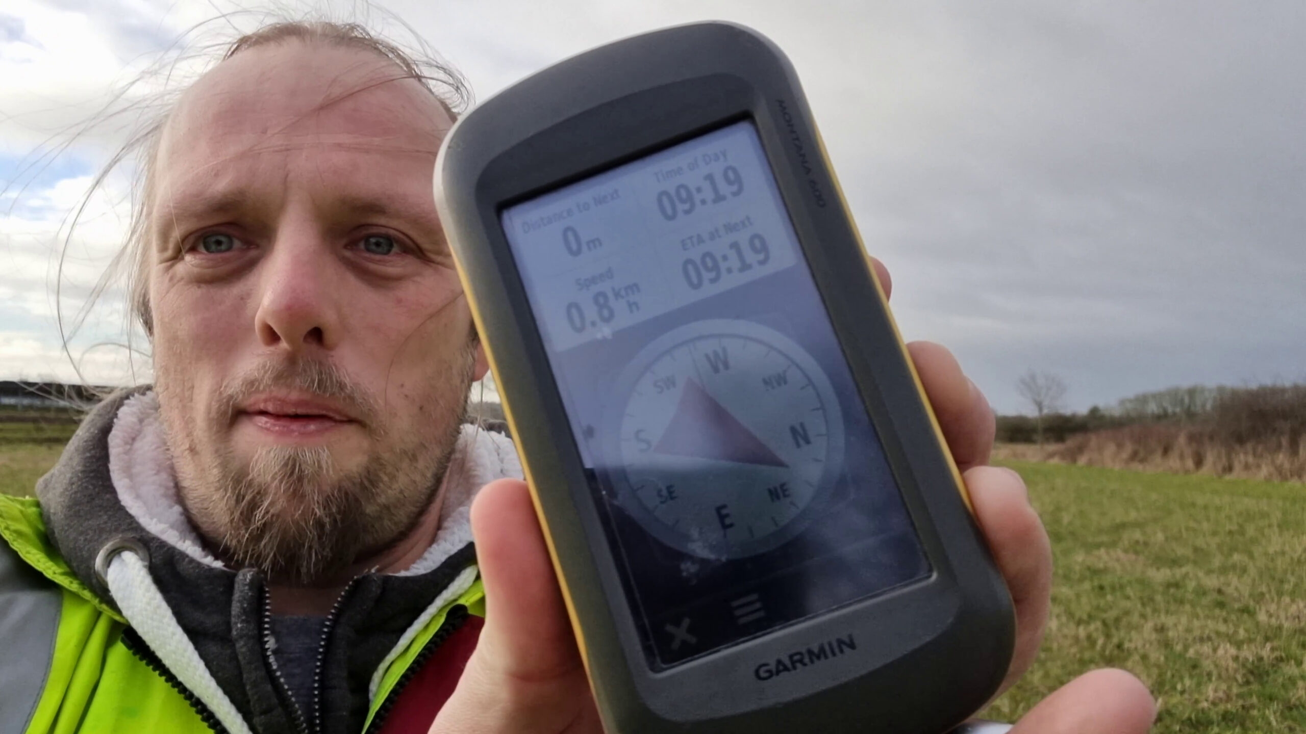 Dan, outdoors in a field on a grey day and with the wind whipping his hair across his face, wearing a high-vis jacket over a warm fleece, holds up a GPS receiver which shows he's zero metres from his destination.
