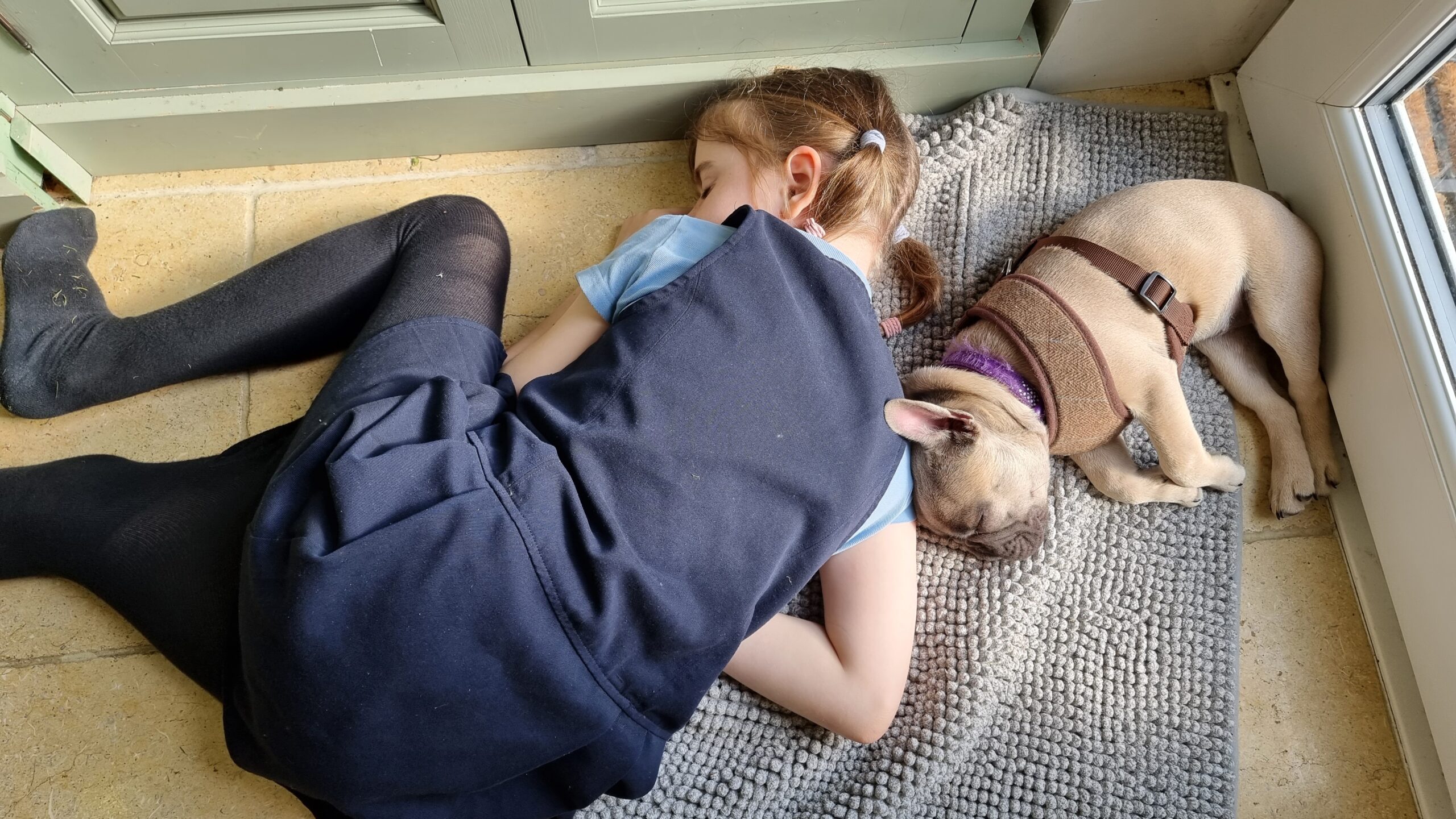 A young girl asleep on a stone floor, her head on a doormat, napping alongside a French Bulldog.