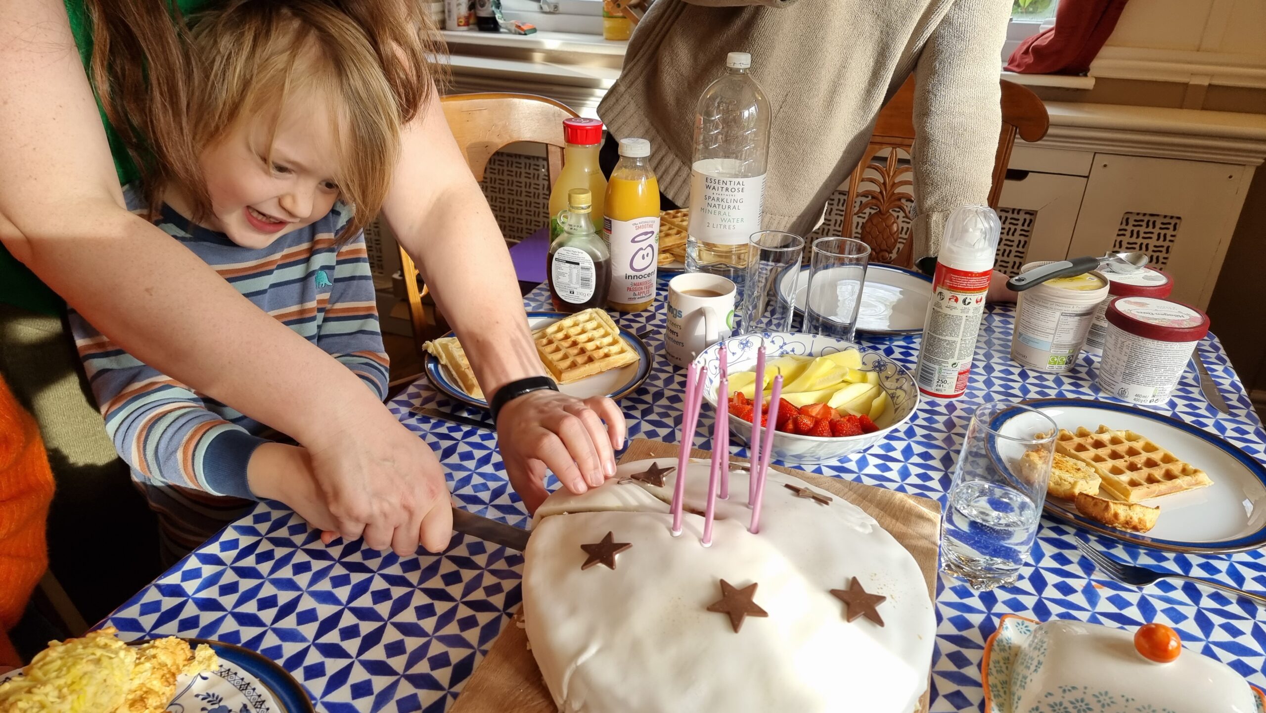 A boy in blue-and-brown striped pyjamas, on a table stacked with plates of waffles, slices into a birthday cake with six candles.