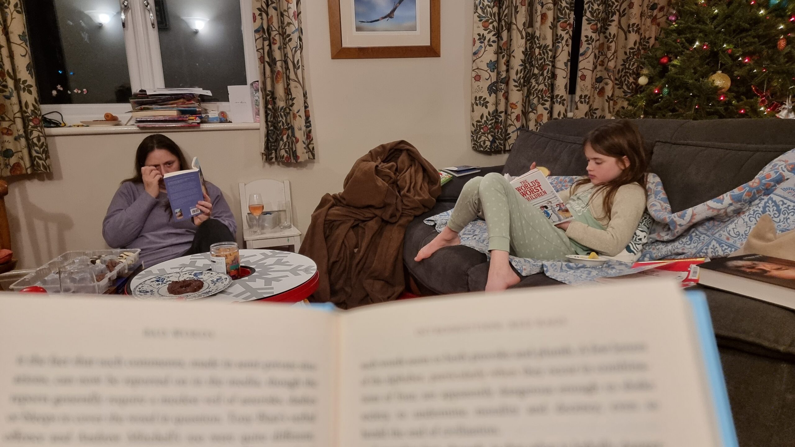 Family members read books in a living room, seen over the top of the pages of an (out of focus) book in the foreground.