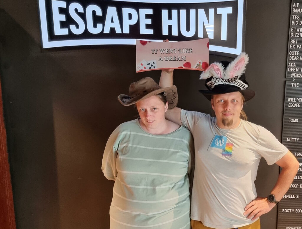 Ruth and Dan hold up an Alice In Wonderland themed sign reading "it went like a dream" underneath the sign for escape room company Escape Hunt. Both are wearing silly hats, and Dan is also wearing white rabbit ears.