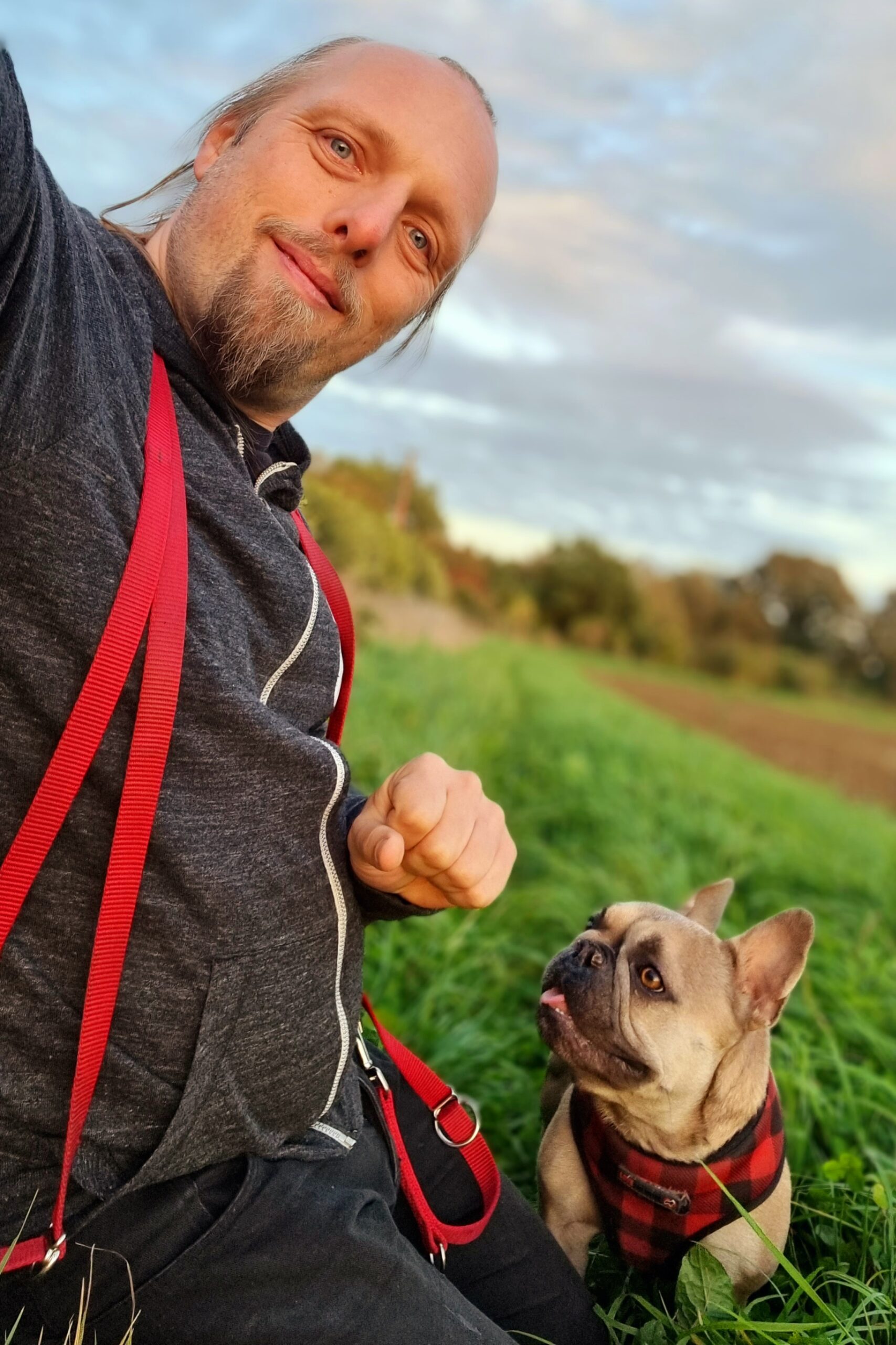 Dan kneels in the grassy verge of a recently-ploughed field in late-afternoon summer sunshine, a long red dog's lead hanging around his shoulders. By his side, a champagne French Bulldog looks with anticipation up at him (or possibly at his closed fist).