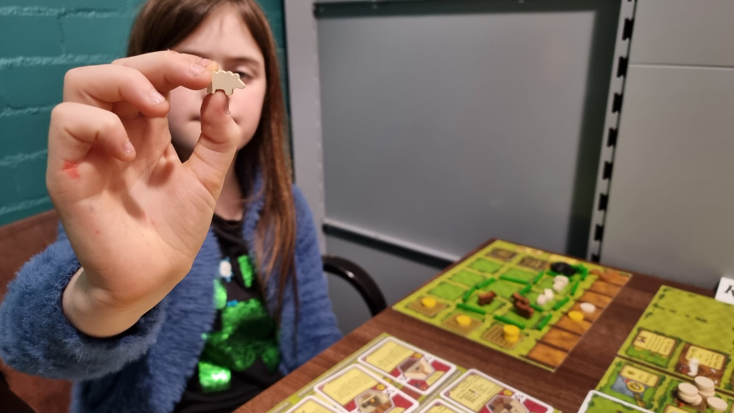 A girl, sat in front of an Agricola farmyard board, holds up a "sheeple" (small wooden sheep game piece) for the camera.