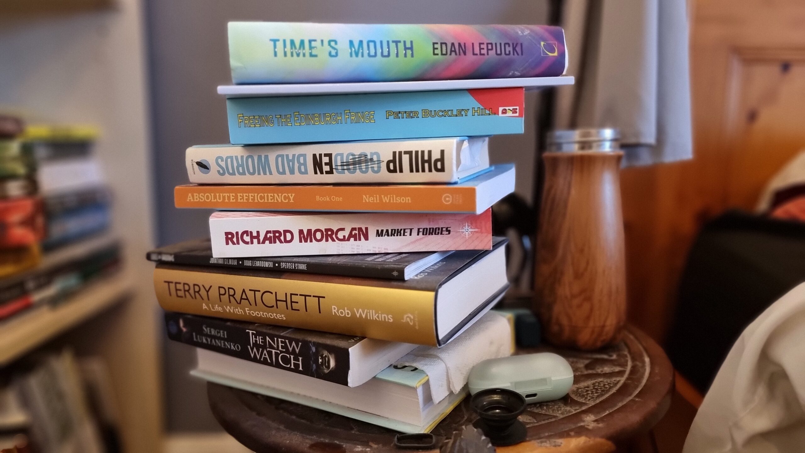 A pile of books on a round-topped wooden bedside table. From top to bottom: Time's Mouth by Edan Lepucki, a slim book whose spine doesn't show a name, Bad Words by Philip Gooden, Absolute Efficiency: Book One by Neil Wilson, Market Forces by Richard Morgan, Kids on Brooms by Jonathan Gilmour, Doug Levandowski, and Spenser Starke, Terry Pratchett: A Life With Footnotes by Rob Wilkins, The New Watch by Sergei Lukyanenko, and a book whose spine is turned away from the camera.