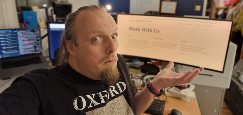Dan, wearing an Oxford-branded t-shirt, shrugs and looks confused in front of a screen showing Automattic's "Work With Us" page.