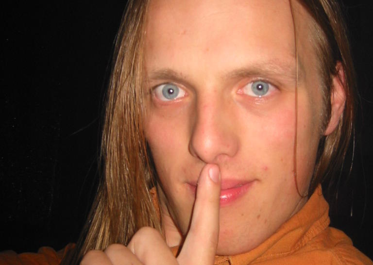 Dan, aged 22, his hair untied and hanging down, wearing an orange shirt, puts his finger to his lips in a "shh" gesture while looking directly into the camera.