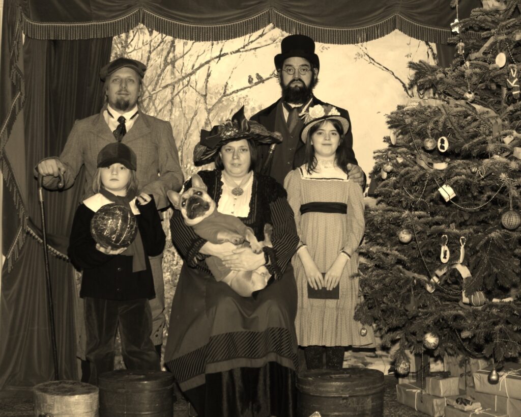 Posed sepia photograph of Dan, JTA, Ruth, and their children and dog, dressed in Victorian-era clothing, by a Christmas tree.