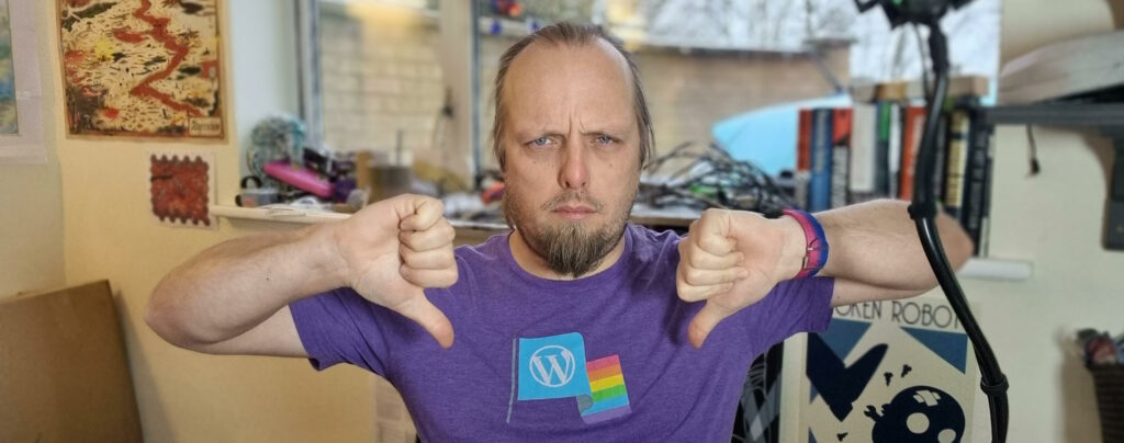 Dan, wearing a purple t-shirt with a WordPress logo and a Pride flag, sits in his home office and gives two "thumbs down" signs while frowning at the camera.