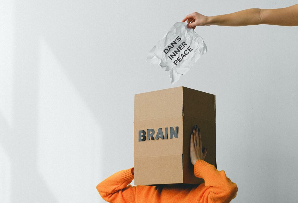A person in an orange jumper holds a cardboard box labelled "BRAIN", which completely covers their head. From outside the image, an hand above the box is holding a piece of paper labelled "DAN'S INNER PEACE".