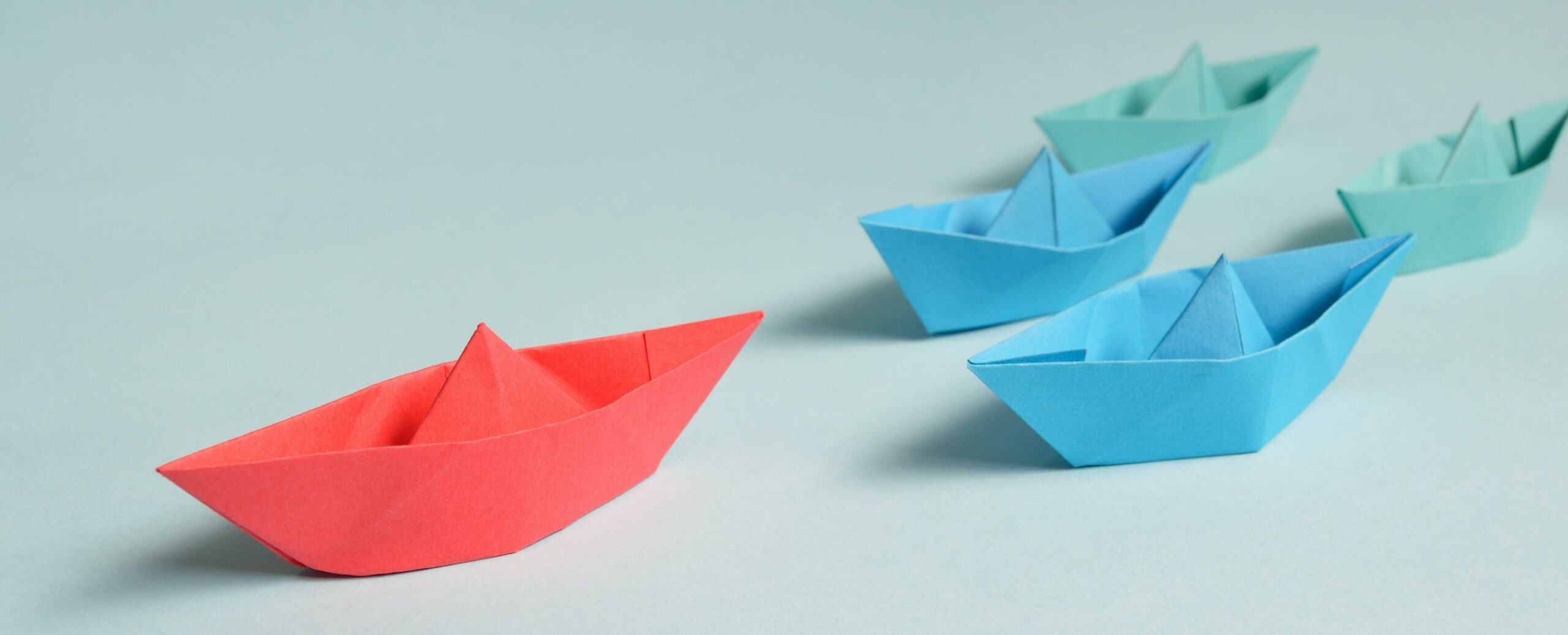 A flotilla of paper boats on a table: a red boat leads the way ahead of two blue and two green boats.