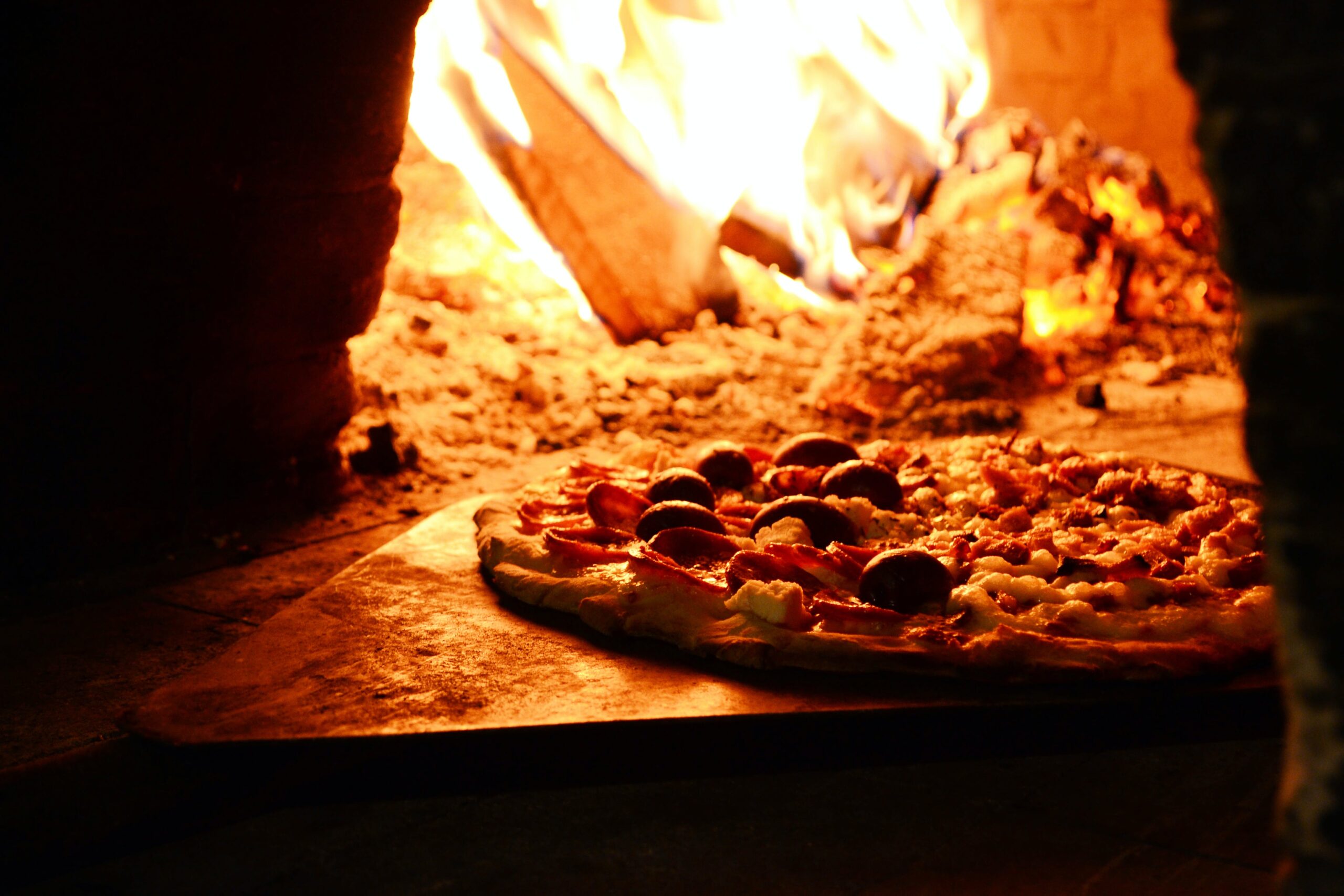 A pizza slides off a peel into a wood-fired oven. One half seems to have salami and olives, the other half perhaps just cheese and tomato.