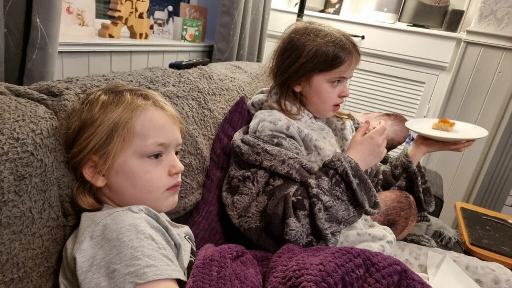 A young boy and a less-young girl sit on a sofa in pyjamas and dressing gowns.