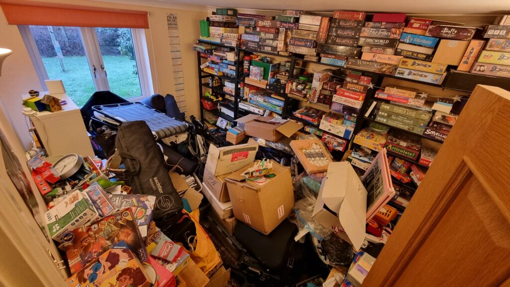 An extremely cluttered room: shelved board games line the walls, heaps of boxes fill all the floor space and are stacked chest-high.