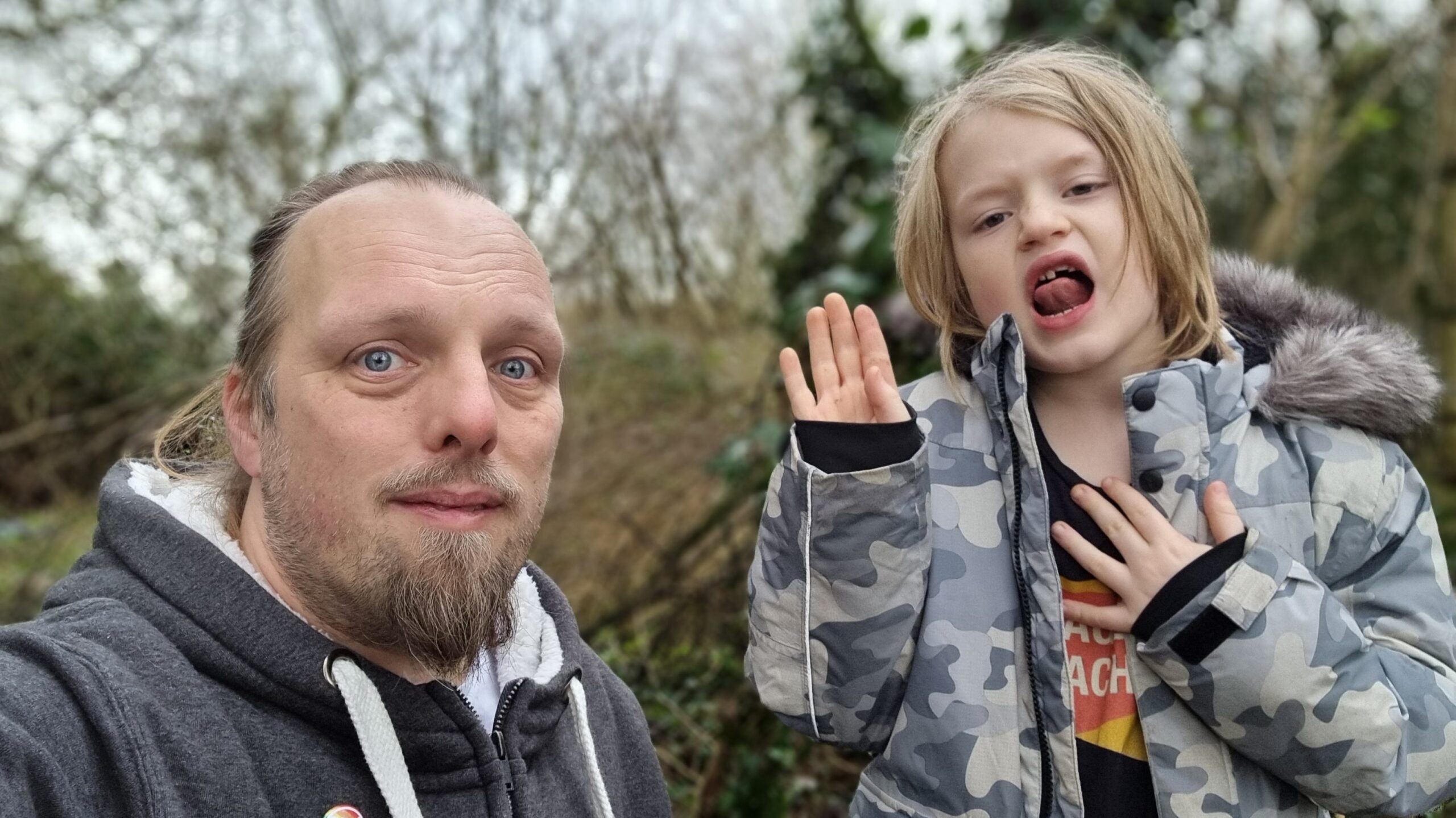 Dan, in a grey jumper, stand beside a 7-year-old boy wearing a camo jacket, in a lightly wooded area.