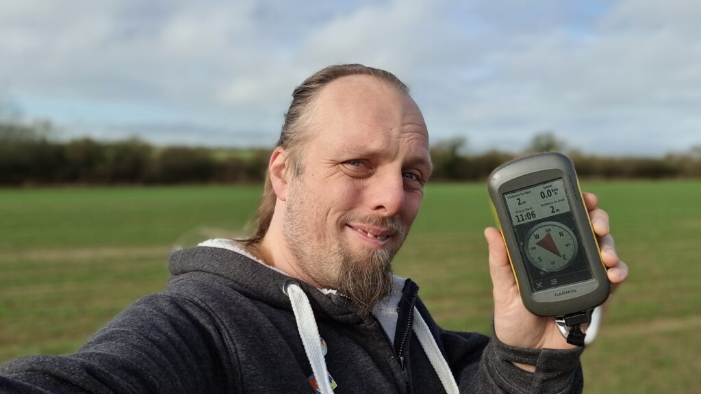 Dan, smiling, holding up his GPSr, in a field.