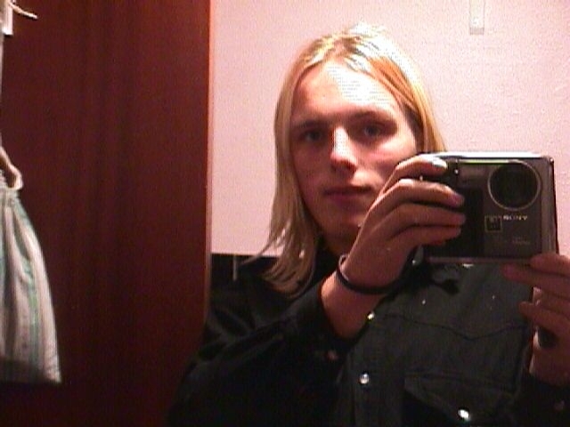 Dan aged 17 - a young white man with platinum blonde shoulder-length hair - stands in front of a pink wall, holding up a large, boxy digital camera.