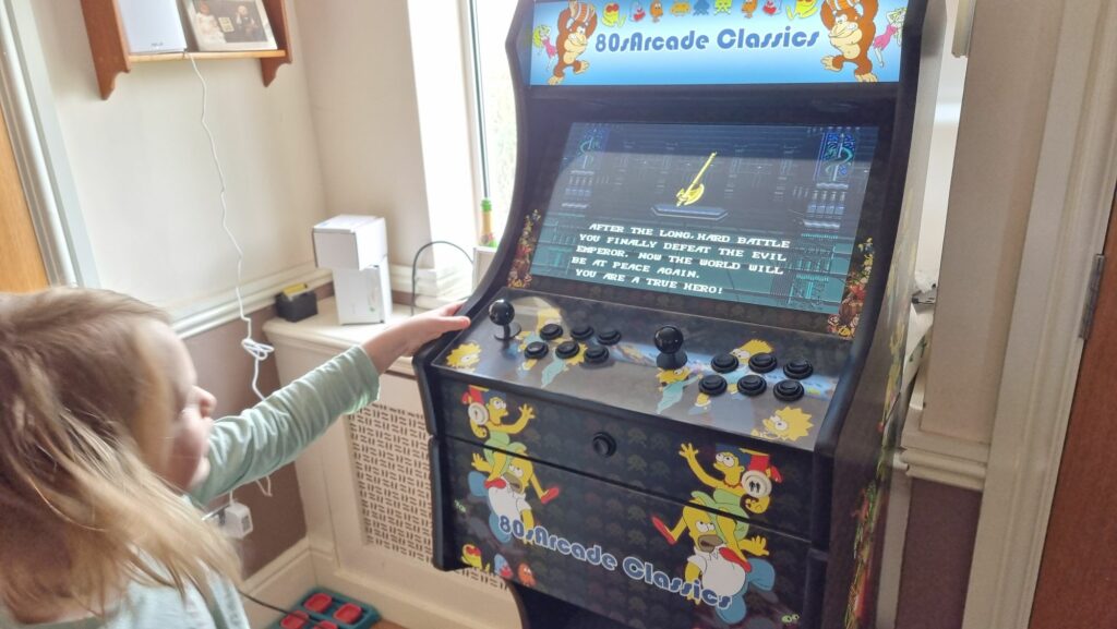 A young boy in green pyjamas stands alongside a video games cabinet. The screen shows 16-bit graphics of a golden axe, below which the text reads: "After a long, hard battle you finally defeat the evil emperor. Now the world will be at peace again. You are a true hero!"