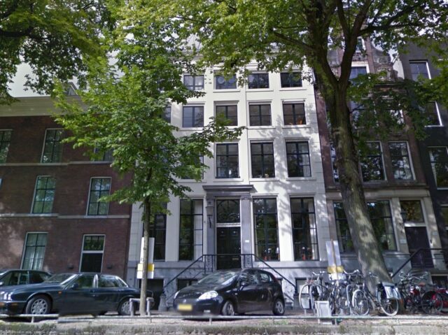 Modern photo, taken from the canal, showing a tall white building in Amsterdam with large windows on the ground floor and also basement level, and an ornamental window above the front door. Photo from Google Street View.