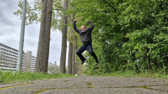 On a tree-lined canalside footpath, Dan leaps theatrically into the air as if in incredible excitement.
