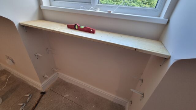 A spirit level on an unfinished shelf, under a window and beneath an uncarpeted floor.