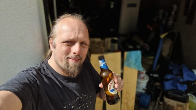 Dan drinks a 0% alcohol beer in front of three upright planks of wood.