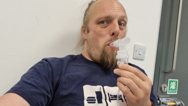 Dan holds a piece of medical apparatus to his mouth.