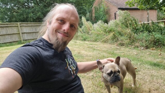 Dan sits on the grass in a garden, with his French Bulldog.