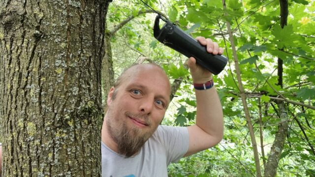 Dan, up a tree, holds a geocache secured high up the trunk.