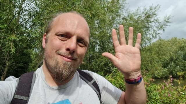 Dan, on an overgrown riverside footpath, waves to the camera.