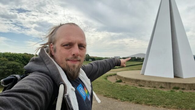 Dan, near a white pyramid-shaped sculpture, points at a distant hill-shaped building.