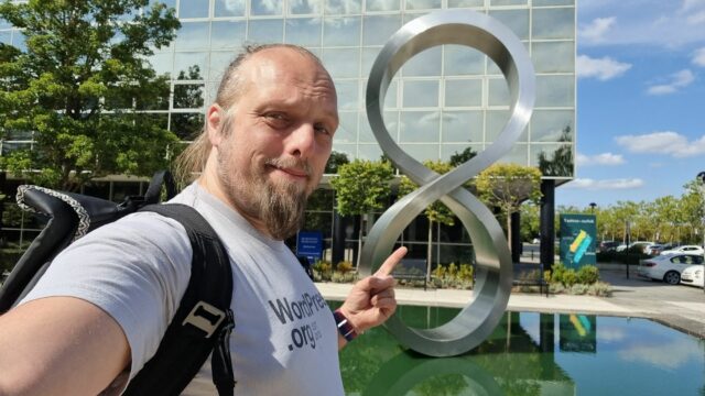 Dan poses for a selfie in front of a figure-8/moebius strip sculpture, on a sunny day.