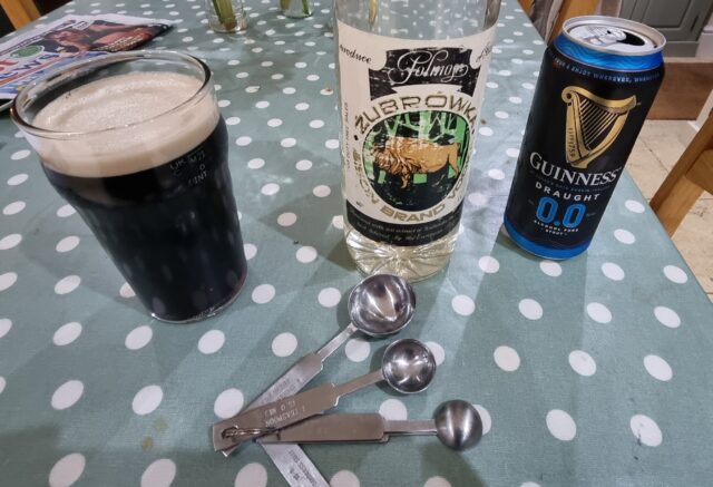 Pint glass having been filled with a carefully measured mixture of 0%ABV Guinness and vodka, with measuring spoons alongside.