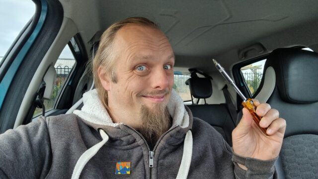 Dan, sitting in a car, holding a screwdriver and smiling.