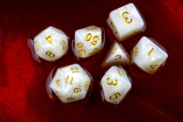 A 'full set' of white polyhedral dice commonly-used by roleplayers - a D4, D6, D8, two D10s, a D12, and a D20 - sit half-submerged in a red liquid.