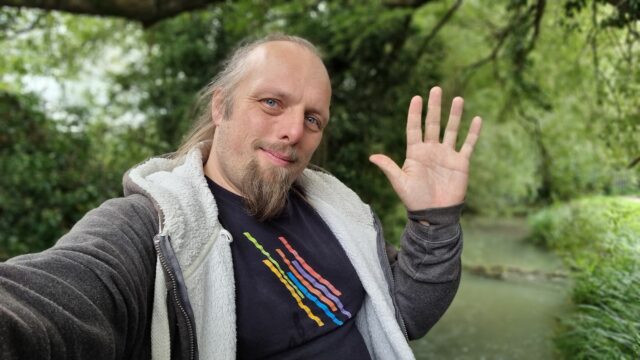 Dan, sitting on a bridge edge near a wooded stream, waves to the camera. 