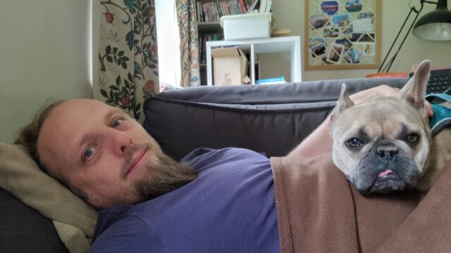 Dan, lying on a sofa under a brown blanket, on top of which is a contented-looking French Bulldog.