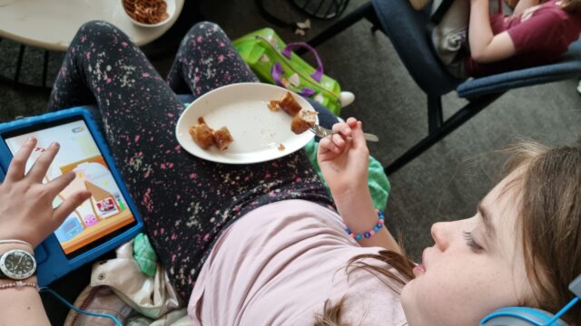 A child slouches in an airport lounge chair. Her right hand is being used to eat a plate of sausages. Her left hand is playing 'Fork N Sausage' on a tablet.