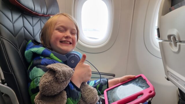 A boy smiles widely as he sits, playing with his tablet, in an aeroplane window seat.