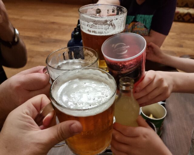 Three glasses of beer held by adult hands clink together against a glass of water and a bottle of Fanta held 6 by cold hands.