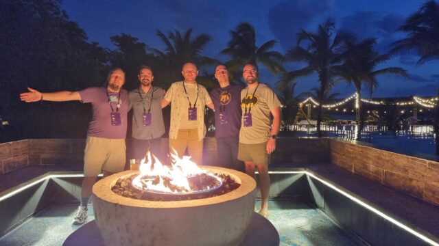 Dan and four other men stand around a firepit, in front of a tropical beach and a twilight sky.