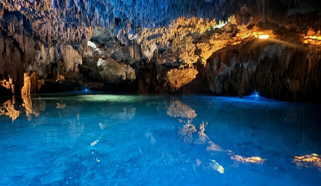 Stalactite-strewn cave deeply filled with clear blue water.