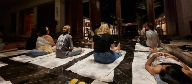 A group of people lie or sit cross-legged on towels in front of a collection of musical instruments.