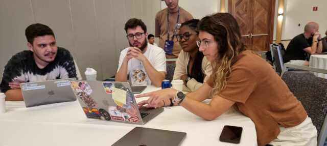 Two men and two women sit around a sticker-covered Macbook laptop, collectively looking at its screen.