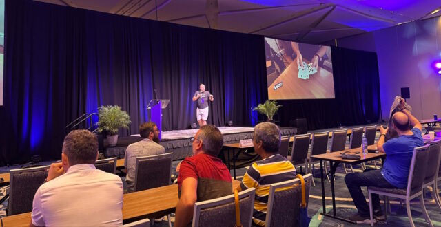 Dan standing on stage in front of a seated audience; a screen behind him shows a close-up of his hands holding several playing cards.