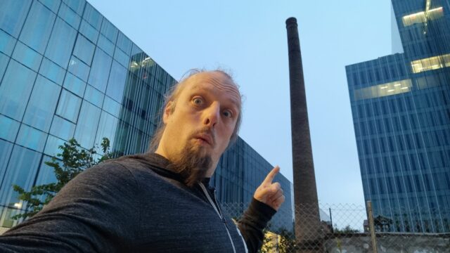 Dan points at an old brick chimney, standing alone among modern glass office buildings.