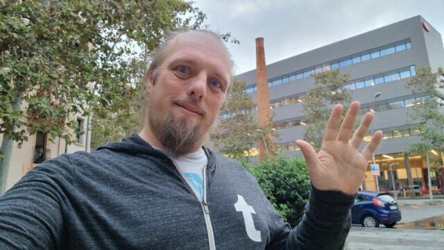 Dan waves in front of an old brick chimney surrounded by modern office and retail buildings.