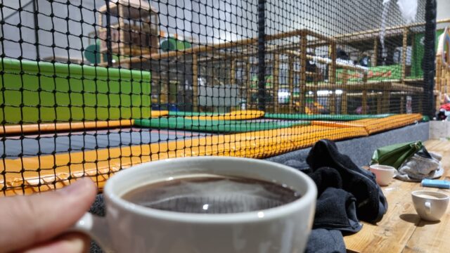 A mug of coffee held in front of a view of a multicoloured soft playground.