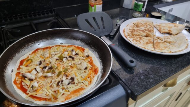 Gas stovetop.a frying pan contains a tortilla wrap topped with tomato sauce, cheese, mushrooms, and jalapeños. Beside its a plate containing a completed quesapizza: two crispy tortilla wraps sandwiching their contents.