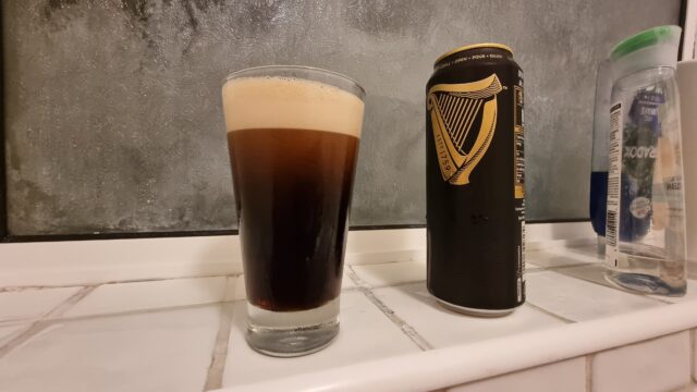 A pint of Guinness alongside a can, on a tiled bathroom shelf.