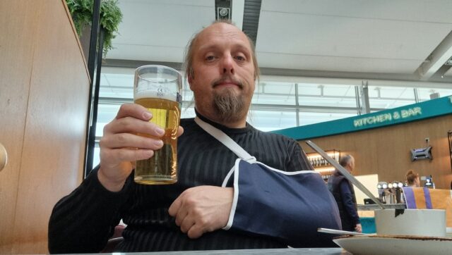 Dan sits in an airport bar drinking a beer held in his right hand. His left arm is in a sling.